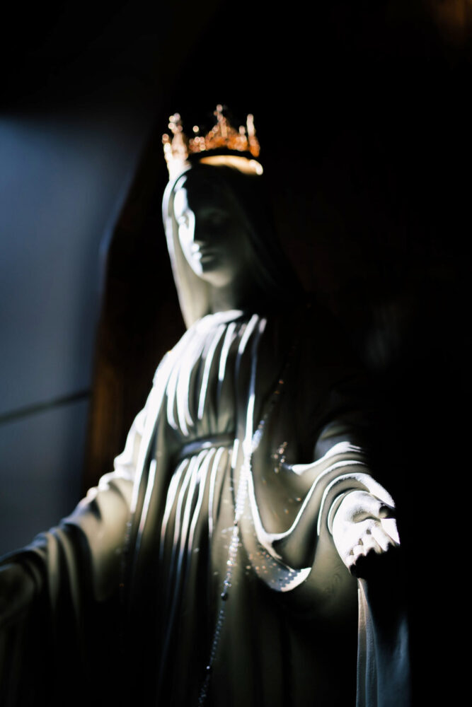 White statue of Mother Mary / Mother Divine with palms up and a gold crown on her head.