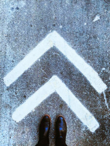 A pair of shoes standing in front of two white forward pointing chevrons that are indicating this is the way.