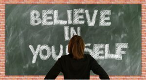 Woman confidently standing facing a blackboard that says in bold caps: Believe in Yourself