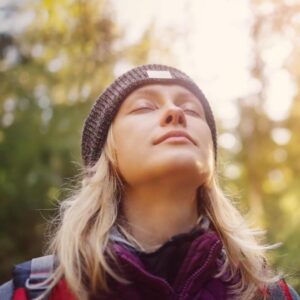 A young woman with closed eyes stands peacefully in a sunlit forest, wearing a knit beanie and layered outdoor clothing, basking in the warmth and serenity of nature.