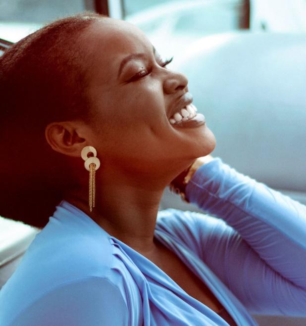 A joyful woman smiling with her eyes closed, wearing a light blue top and statement earrings, radiating confidence and inner peace. She's connecting with her inner wisdom in a powerful way.