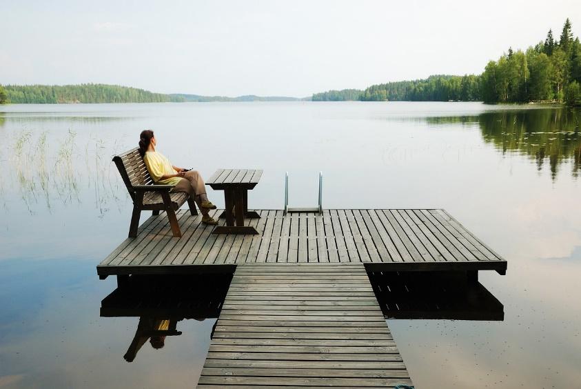 A person sitting alone on a wooden dock overlooking a calm lake surrounded by forest, peacefully gazing into the distance on a quiet day. She could be waiting patiently for the opportunity to take the next step on her spiritual journey.