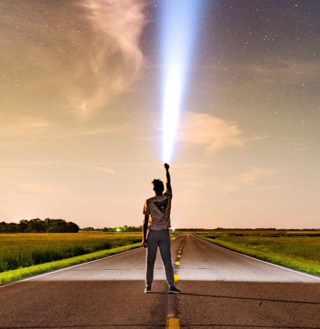 A person standing in the middle of an empty road at night under a starry sky, reaching up to energetically connect with a powerful bright beam of Light.