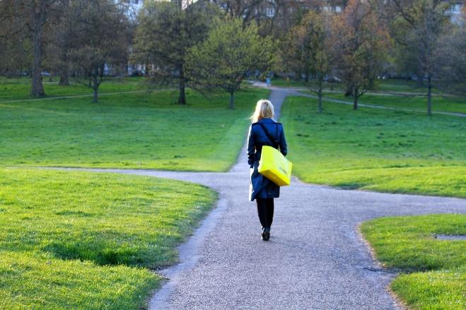 A solitary woman walks briskly along through a grassy green park carrying a large yellow bag and wearing a dark navy coat. She is approaching three diverging paths and confidently chooses the best one for her at this time.