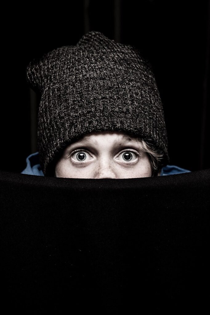 Young child with big eyes peeping over a barrier - looking surprised / afraid / concerned - some strong emotion. Image has a black background so all you can really see are eyes and a pulled down knit hat.