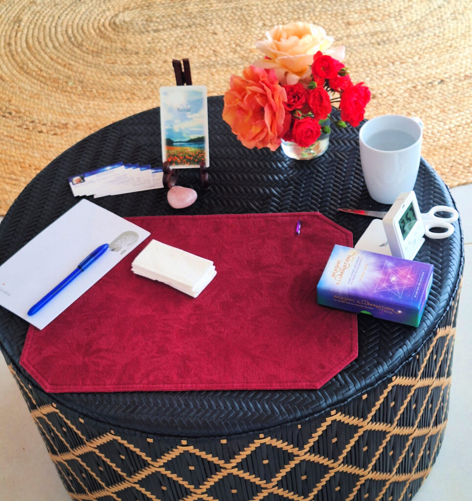 Photo of a small round table set up as an altar with a notebook, Akashic Illumination Deck with one card displayed, cup of water, and a vase of beautiful roses that the story is about.